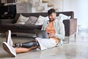 Happy young man with a leg prosthesis sitting on the floor at home using a laptop, representing daily life with an amputation and the potential role of online resources for phantom limb syndrome pain management