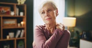 Elderly woman at home experiencing neck pain and shoulder discomfort from joint inflammation, muscle tension, or fibromyalgia-related fatigue.